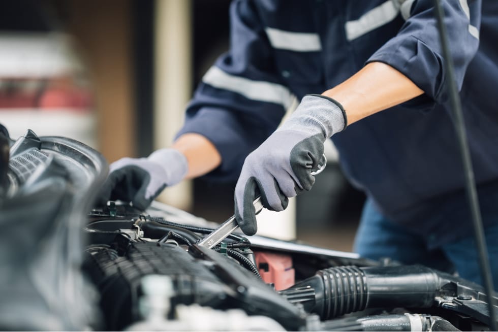 A mechanic wearing grey work gloves uses a combination wrench to work inside the open engine bay of a vehicle.