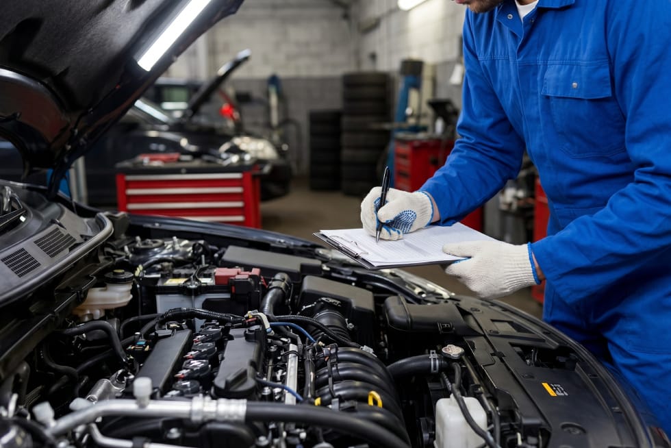 A mechanic in blue coveralls holding a pen and clipboard, filling out a checklist while standing in front of an open car engine bay.