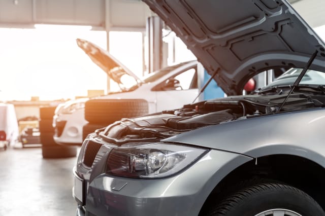 Side angle of a silver BMW with its hood open for inspection alongside other vehicles inside a bright auto repair garage.