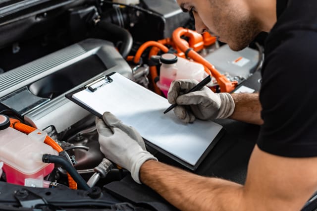 Close-up of an auto mechanic filling out a diagnostic checklist on a clipboard while inspecting an open vehicle engine bay.